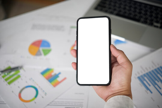 Close up hand holding mobile phone mockup isolated white screen with laptop background. Digital marketing and online banking concept. Professional suit worker using smartphone.