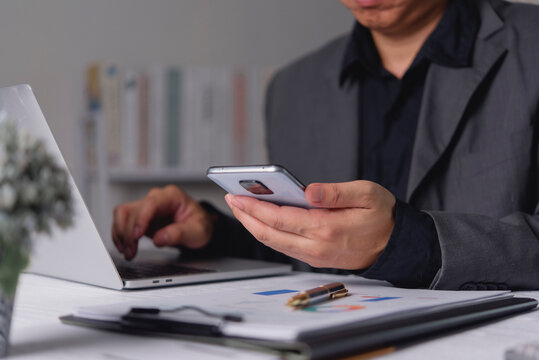 Businessman hand holding smartphone typing laptop computer desk blurred financial chart corporate office multitasking digital communication cross device workflow synchronization efficiency.