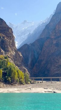 The video is from a boat sailing on Attabad Lake, Hunza, Pakistan. The backdrop features snow-capped mountains, poplar trees with yellow leaves, and the sparkling blue water on a sunny day.