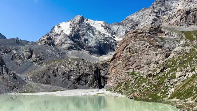 The lake with a snow-covered mountain view on a rocky slope in the Tianshan Mountains, Xinjiang, China. There is a waterfall flowing from the peak on a sunny day with a blue sky. 