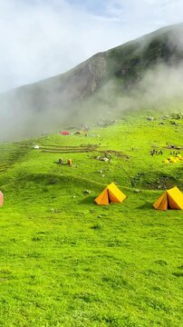 Panning video shows many hikers standing on a green grassy hillside campsite at Bhrigu Lake Trek. There were several colorful tents on the lawn with small flowers on a cloudy day.