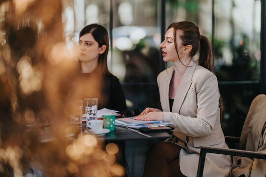 Two professional women sit at a cafe table reviewing documents and talking. The colleagues continue their business discussion over coffee and paperwork in a relaxed setting.