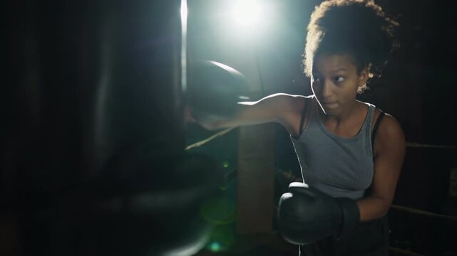 Young african woman fighting with punching bag, cinematic portrait in gym. Professional female boxer fighter training alone in dark boxing ring, release aggression and negative emotions, do sport