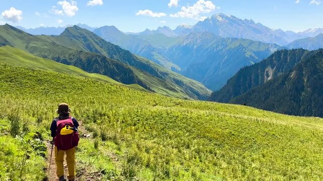 Wide-angle view of green grassy hills on both sides at Tianshan Mountain, Xinjiang, China. A woman walking in the distance on a sunny day. Many mountains are forming beautiful layers.