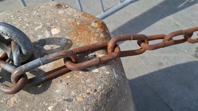 Close-up of heavy rusty metal chain secured to a concrete pillar with a steel eye bolt. Industrial detail represents security, strength, and old urban infrastructure in a city environment