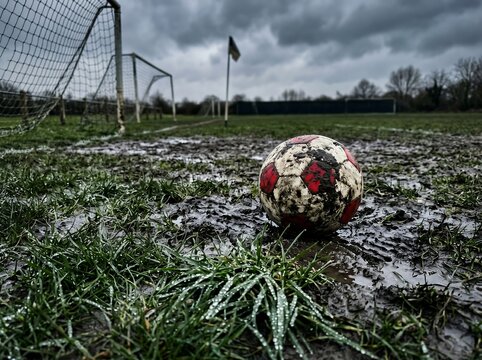Muddy soccer ball on wet grass field with goal net in background on a cloudy day