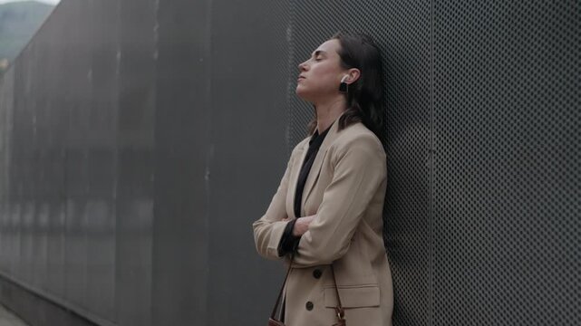 Calm woman with eyes closed enjoying music on wireless earbuds while leaning against a city wall.