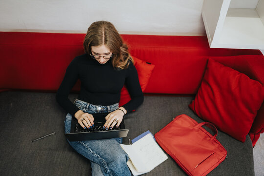 A student sits on a red couch typing on a laptop while taking notes. The young woman studies at home with a notebook and backpack beside her.