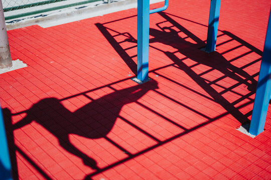 A child climbing monkey bars casts a strong silhouette on red playground flooring. The playground equipment and shadow create bold shapes and contrast under bright sunlight.