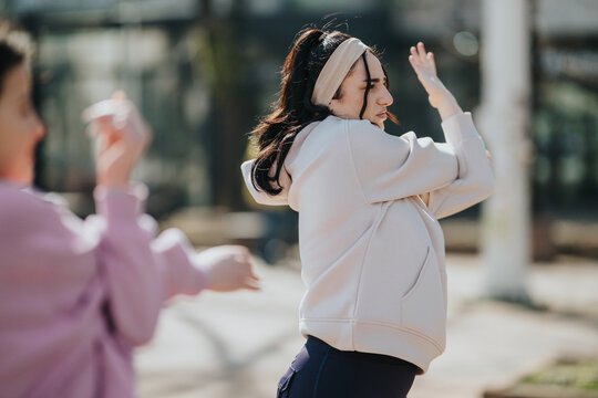 Two women stretch and warm up in an outdoor setting while wearing casual active wear. They perform side stretches together before exercising in a park-like urban area.