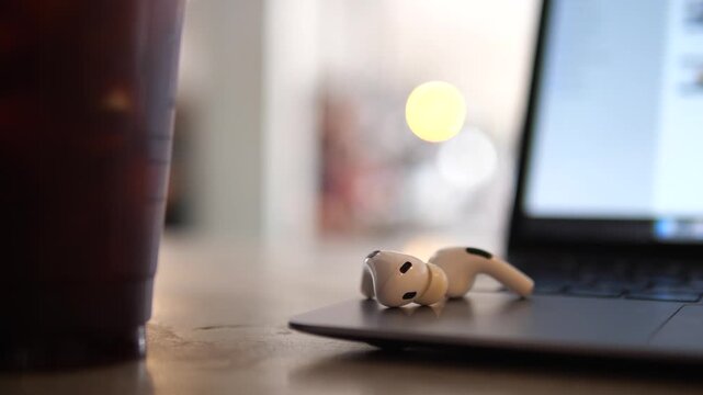 Cinematic close-up of white wireless earbuds on a laptop keyboard with a glass of iced coffee in a blurred background, modern remote workspace and cafe lifestyle concept