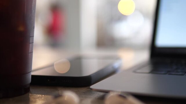 Cinematic close-up of white wireless earbuds on a cafe table with a blurred smartphone, laptop, and iced coffee in the background, modern remote workspace concept