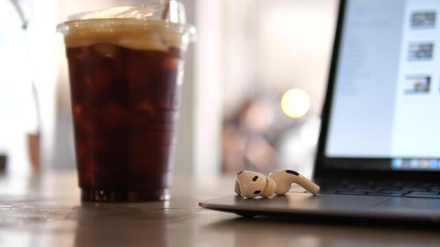 inematic close-up of white wireless earbuds on a laptop keyboard with a glass of iced coffee in a blurred background, modern remote workspace and cafe lifestyle concept