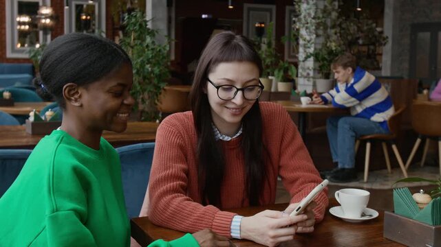 Multiethnic young girl friends looking at smartphone and having fun conversation while dining out