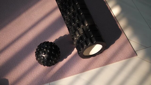 Cinematic close-up of a black textured foam roller and massage ball on a pink yoga mat with beautiful window blind shadows, home fitness and muscle recovery concept