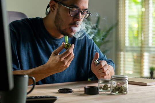 Man smoking a cannabis from smoking pipe, with cannabis equipment and accessories on the table.