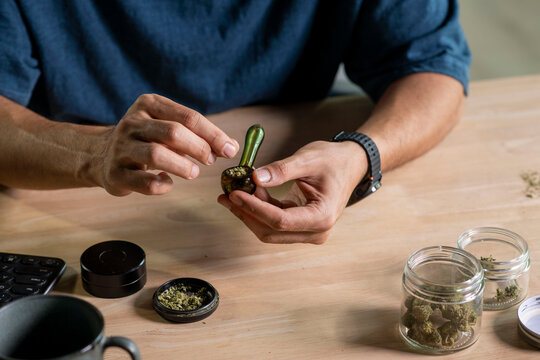 Man filling glass pipe with cannabis, with cannabis equipment and accessories on the table.
