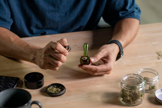 Man smoking a cannabis from smoking pipe, with cannabis equipment and accessories on the table.