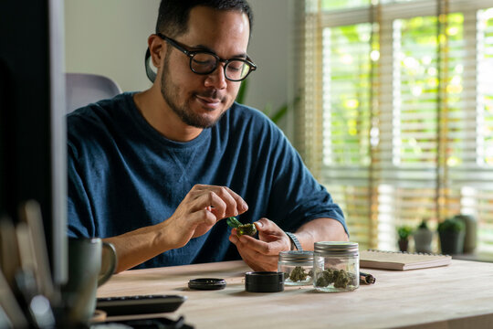 Man filling glass pipe with cannabis, with cannabis equipment and accessories on the table.