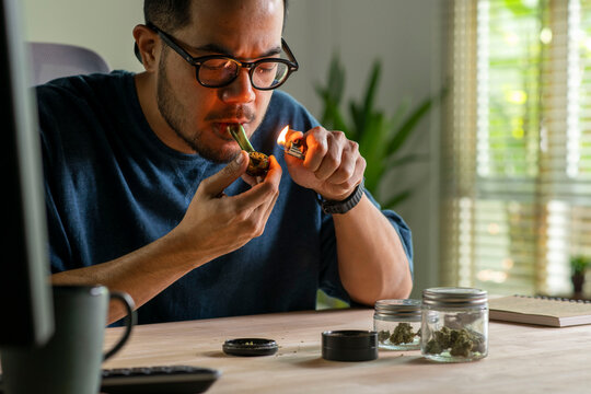 Man smoking a cannabis from smoking pipe, with cannabis equipment and accessories on the table.