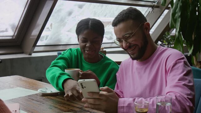 Cheerful Black girl with coffee cup and multiethnic young man looking at smartphone discussing photo at restaurant table