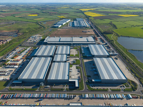 Aerial view of the iPort logistics park featuring large modern warehouses, loading docks, and rows of parked trailers surrounded by green fields and a small lake in Loversall, England, United Kingdom.