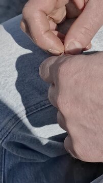Hands of an individual focused on a small task, holding a tiny paper scrap against a backdrop of blue jeans. Bright daylight emphasizes an authentic moment of checking a hidden message or a list