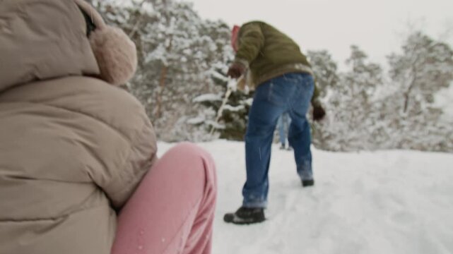 Guy pulling red sled with young woman uphill, man helping him by pushing it while group of friends enjoying winter activity outdoors