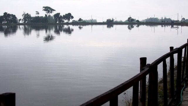 View of calm Siman Reservoir on a cloudy afternoon from behind a wooden boundary fence, featuring still water reflecting distant trees in the rural landscape of Kediri, Indonesia, Southeast Asia.