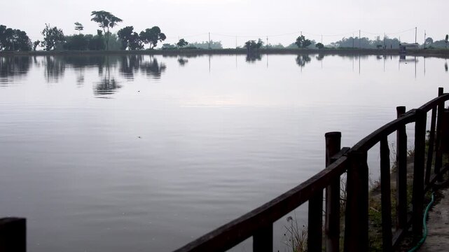 Panning shot of the calm Siman Reservoir on an overcast afternoon from behind a wooden boundary fence, showing still water and distant trees in Kediri, Indonesia, Southeast Asia.