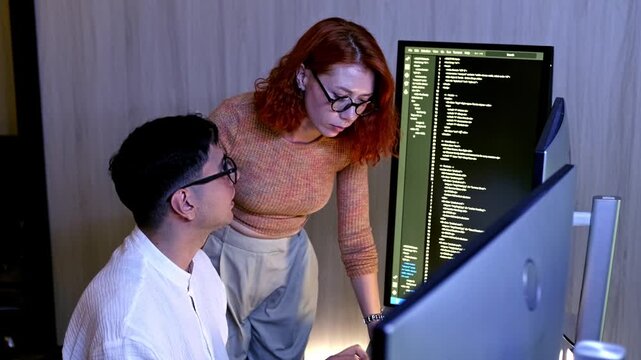 Two programmers collaborate at a dual-monitor workstation, reviewing and debugging code on screen in a modern office.