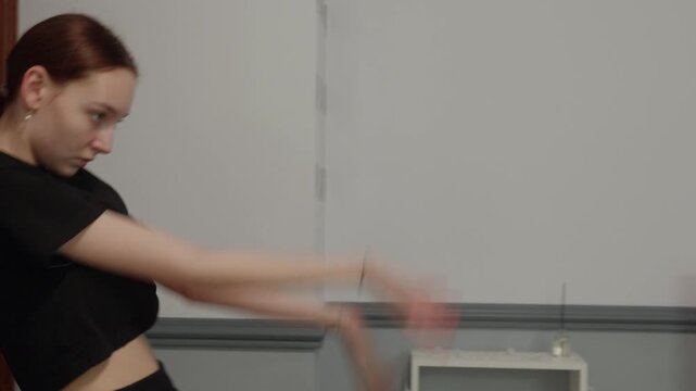Graceful young woman with red hair in black top performs contemporary, modern dance in minimalist room. She moves fluidly, expressing herself through movement