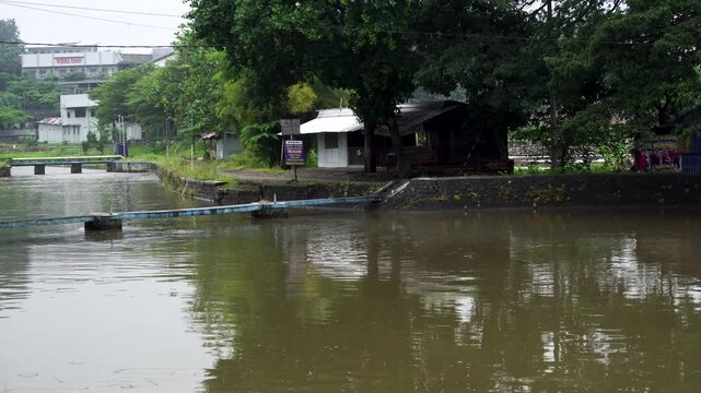 Panning shot following a murky brown river flowing into the Siman reservoir under an overcast afternoon sky in Kediri, Indonesia, Southeast Asia.