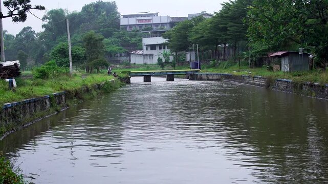 Murky river water flowing towards Siman Reservoir in Kediri, Indonesia. Afternoon view of an irrigation canal with a small bridge, buildings, and green trees along the bank in Southeast Asia.