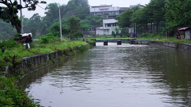 Panning view of a murky river canal flowing into Siman Reservoir, Kediri, Indonesia. Locals ride motorcycles across a background bridge during a cloudy Southeast Asian afternoon.