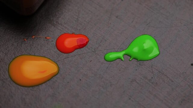 Close-up of vibrant paint droplets in orange, green, and yellow on a textured surface with a brush applying green paint.