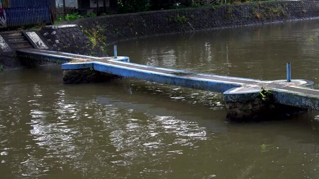 Close up of a small weathered concrete bridge crossing a flowing river stream leading to Siman Reservoir in Kediri, Indonesia, showcasing rural water infrastructure and natural landscape.