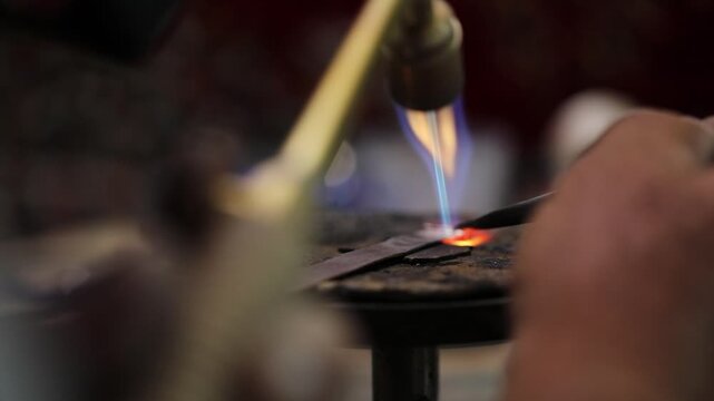 A close-up of a jeweler using a blowtorch to heat a metal piece, showcasing the craftsmanship of metalworking with glowing embers and focused flame.