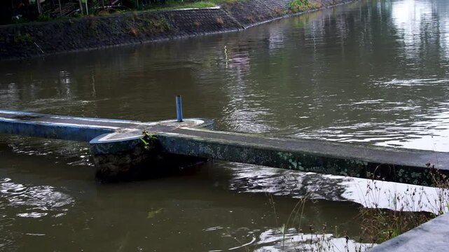 Panning closeup of a small concrete bridge crossing a flowing river canal stream leading to Siman Reservoir in Kediri, Indonesia.
