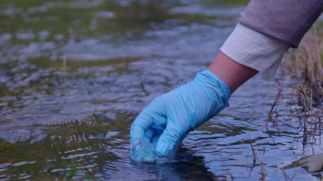Female scientist wearing blue protective gloves collecting a water sample from a stream in a test tube to measure pollution levels and water quality, an important part of environmental analysis
