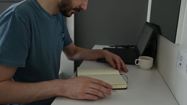 Caucasian man with a beard sitting at a modern desk writing down his thoughts, ideas, or to do list in a notebook, then putting his pen down and closing the diary to finish his task