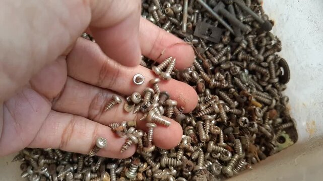 A hand carefully selects and picks up multiple small metal screws from a large messy pile of various industrial fasteners, bolts, and rusty hardware pieces.