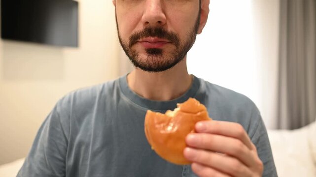 Unrecognizable man with a beard enjoying a delicious fast food meal, biting into a juicy burger and eating french fries. Unhealthy diet and cheat meal concept in a modern apartment