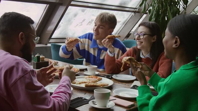 Company of four joyful diverse friends toasting with pizza slices and eating while sitting at table in cafe
