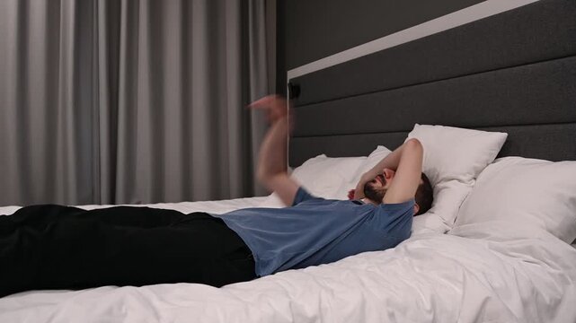 Tired young man collapsing onto a comfortable bed after a long day, falling backwards onto the pillows in a modern hotel room and relaxing before waking up and getting out of bed