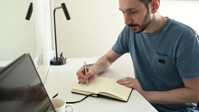 Caucasian man with a beard sitting at a white desk, writing down notes in his journal, finishing his work, and closing the notebook. A male student or employee taking notes in his home office