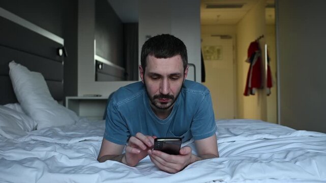 Relaxed young man with a beard lying on a comfortable bed in a hotel room, casually browsing social media, chatting, or watching videos on his smartphone during his leisure time