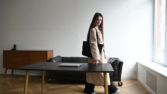 Confident young businesswoman wearing a trench coat and carrying a bag walks into a modern office space, approaching a large window to thoughtfully look at the city view outside