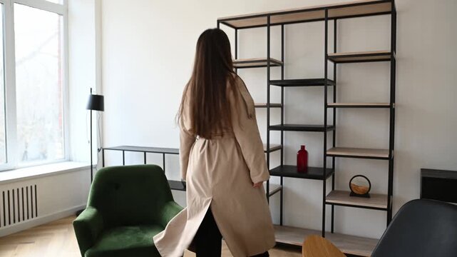 Beautiful young woman wearing a trench coat standing up from a chair and walking around a contemporary and stylishly furnished living room, observing the interior design of the apartment