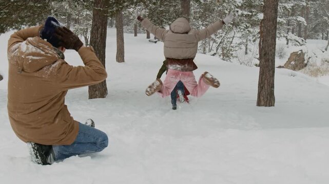 Four cheerful friends having fun outdoors on winter day, girl sledding down snowy slope, friend running after her, man meeting her downhill